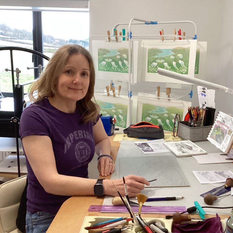 Artist and printmaker Miranda Whitten-Walker sits smiling at her studio desk with a window, etching press and linocut prints on a drying rack behind her.